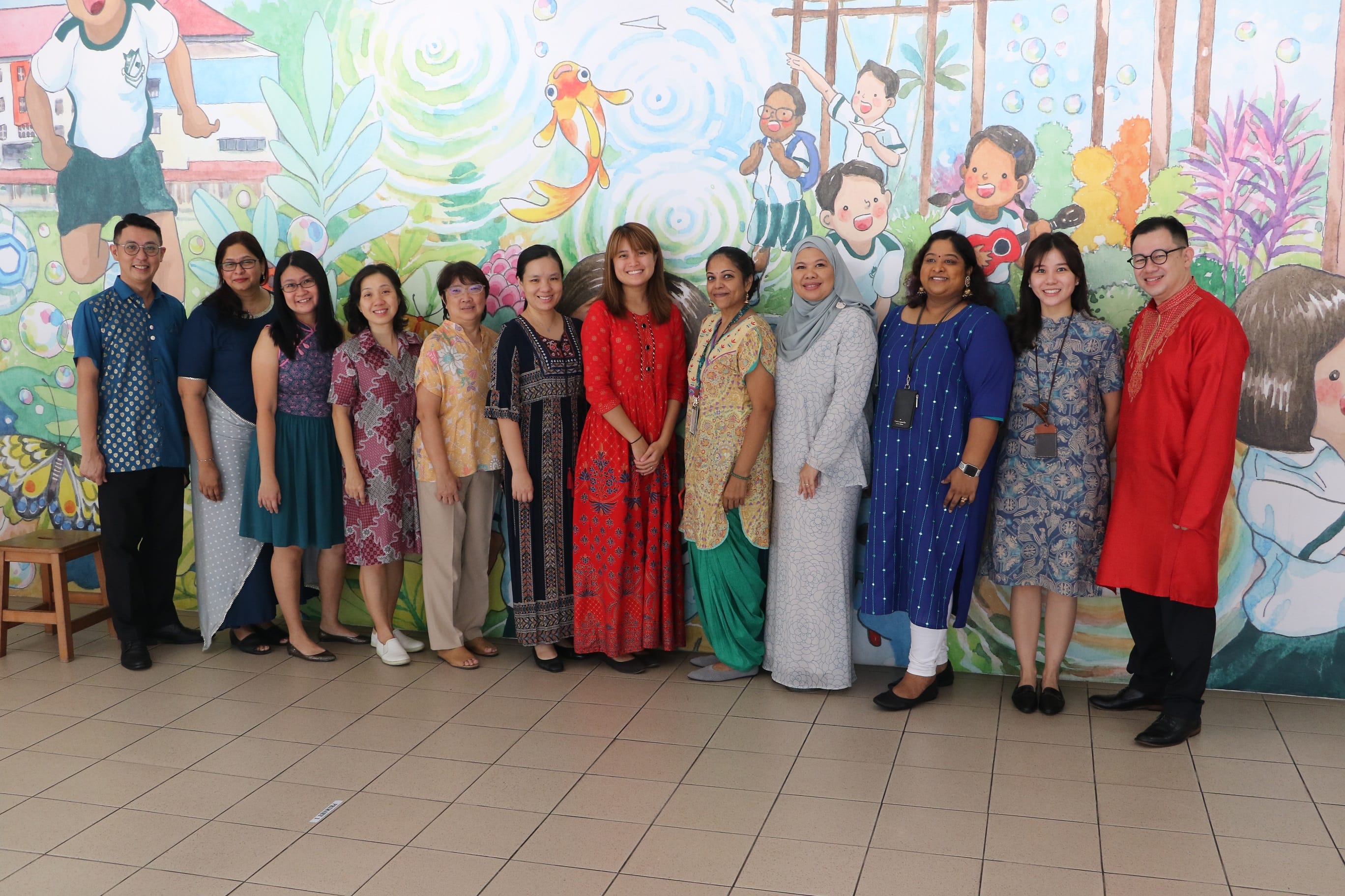 A group of 13 adults in colorful traditional outfits posing together in front of a children's mural.